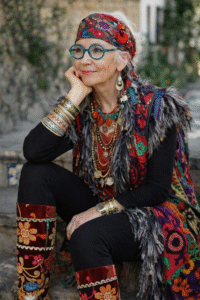 Colorful older woman in layered boho clothing, headscarf, round glasses, and statement jewelry.